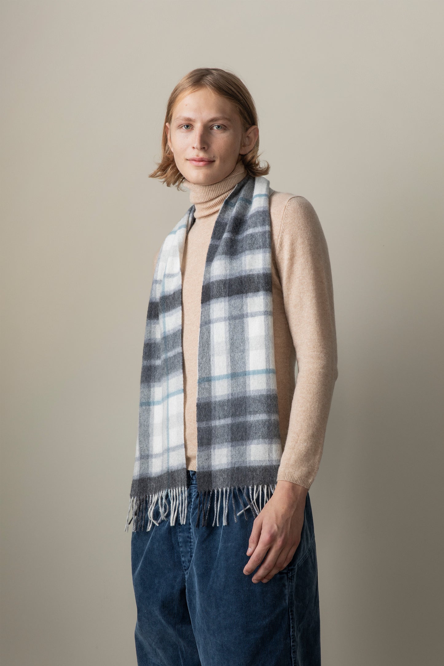 Model wearing Contemporary Check Lambswool Scarf in Ashley Blue draped over a camel turtleneck, showing the plaid pattern and fringed ends
