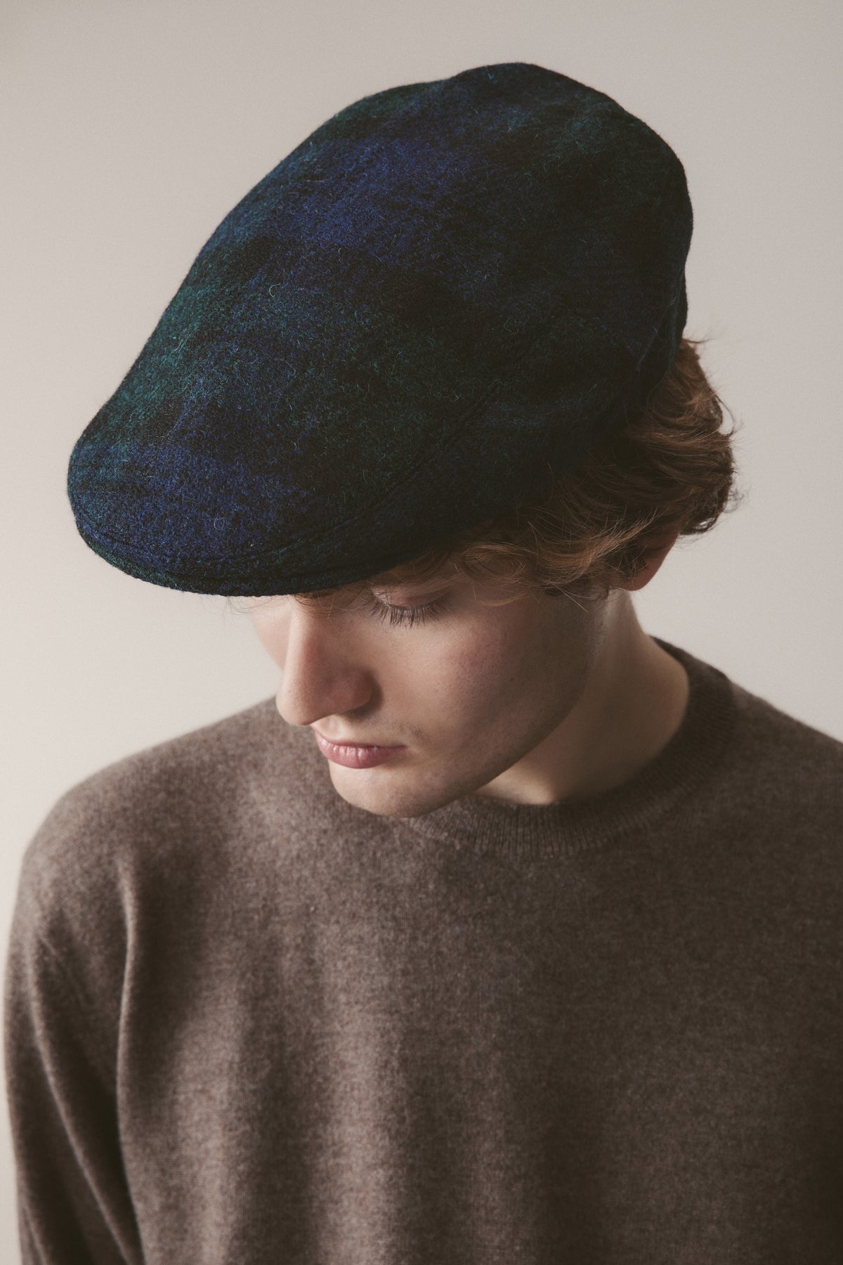 Harris Tweed Stornoway Cap in Blackwatch tartan worn by model looking down, showing the flat cap's profile and wool texture