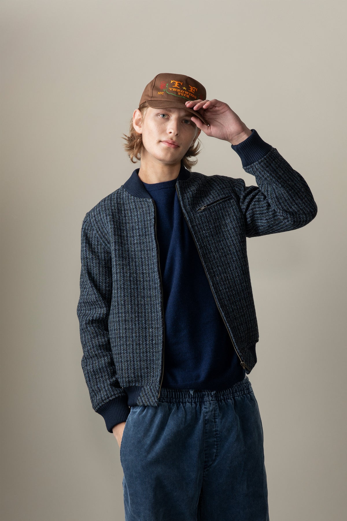 Model wearing Men's Harris Tweed Bomber Jacket in blue dogstooth pattern, unzipped over navy crew neck and corduroy trousers