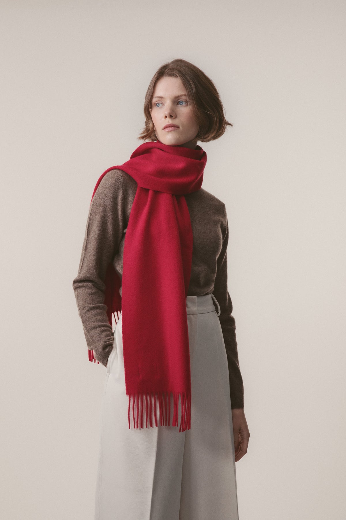 Model wearing the Solid Cashmere Scarf in Red draped around the neck with fringed ends visible, paired with a brown sweater