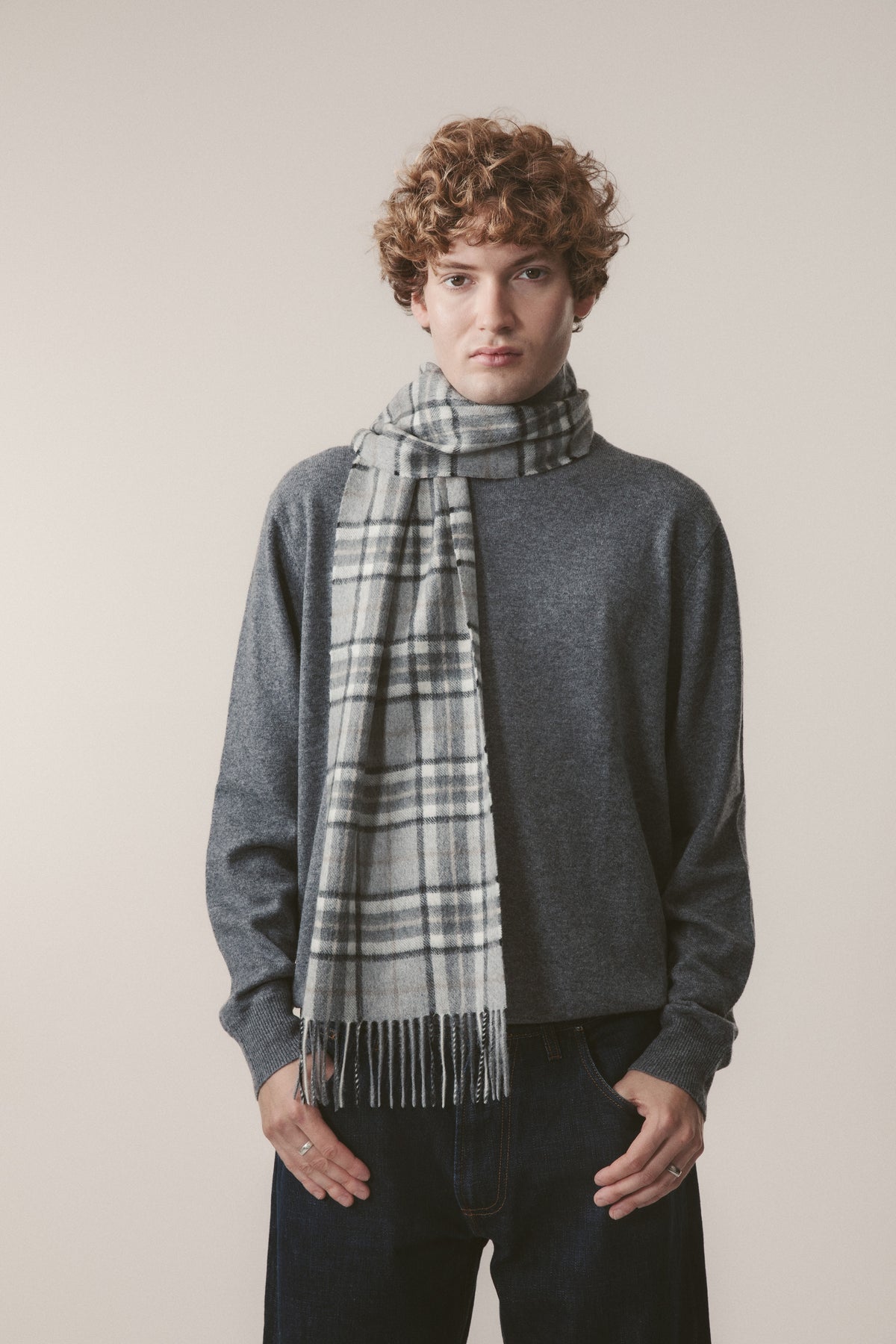 Model wearing the Scotty Thompson Cashmere Scarf in Pewter draped over a grey sweater, showing the plaid pattern and fringe detail
