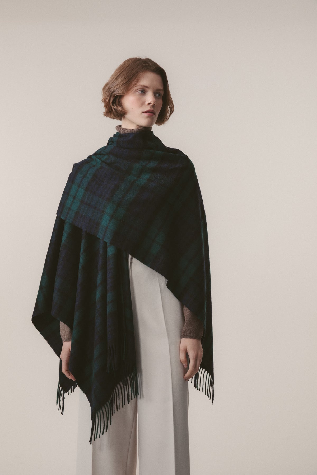 Model wearing Heritage Tartan Cashmere Wrap in Blackwatch draped over shoulders, showing fringed edges and green-navy plaid pattern