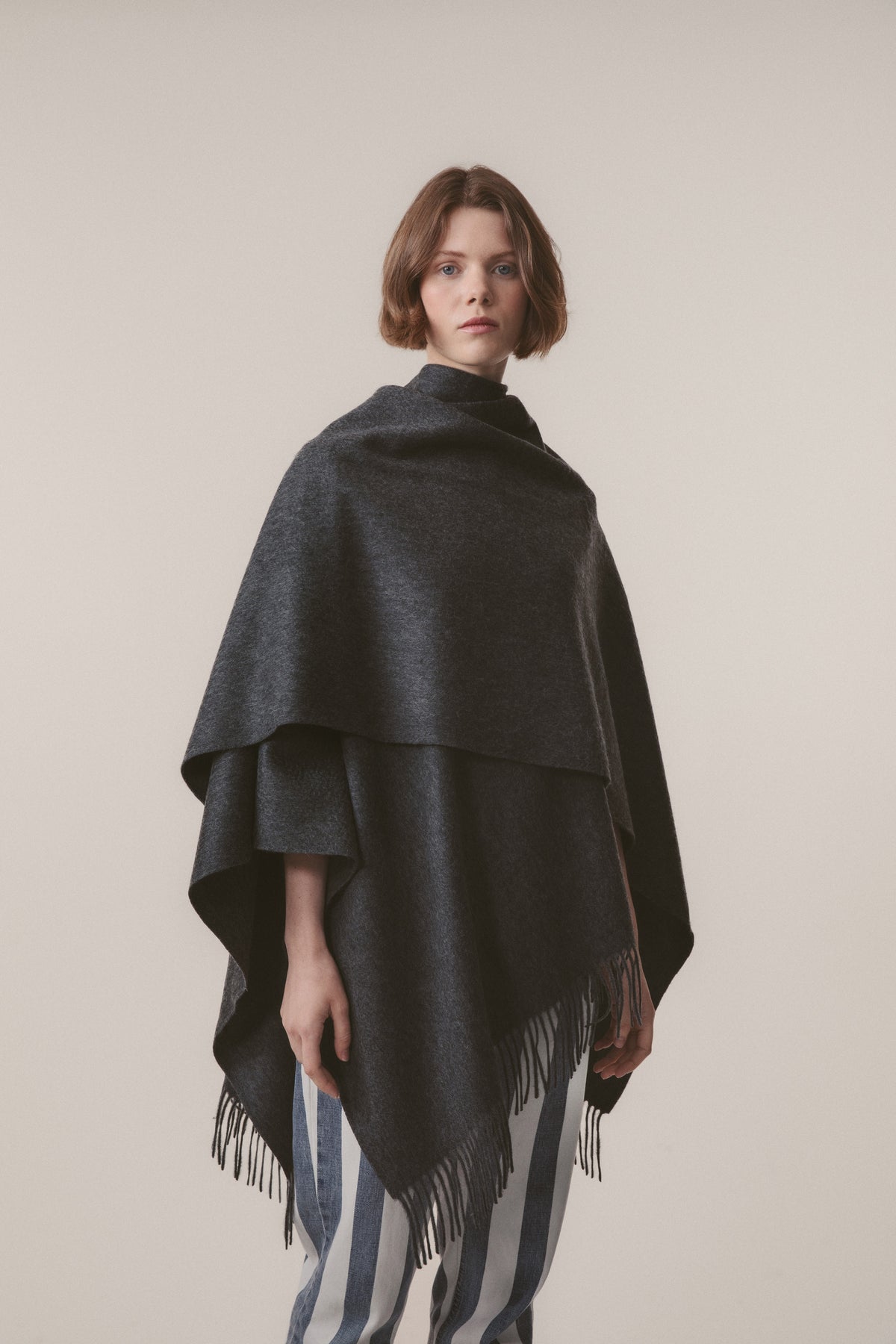 Model wearing Solid Cashmere Wrap in Steinway Grey draped as a poncho, showing fringed hem detail and layered drape