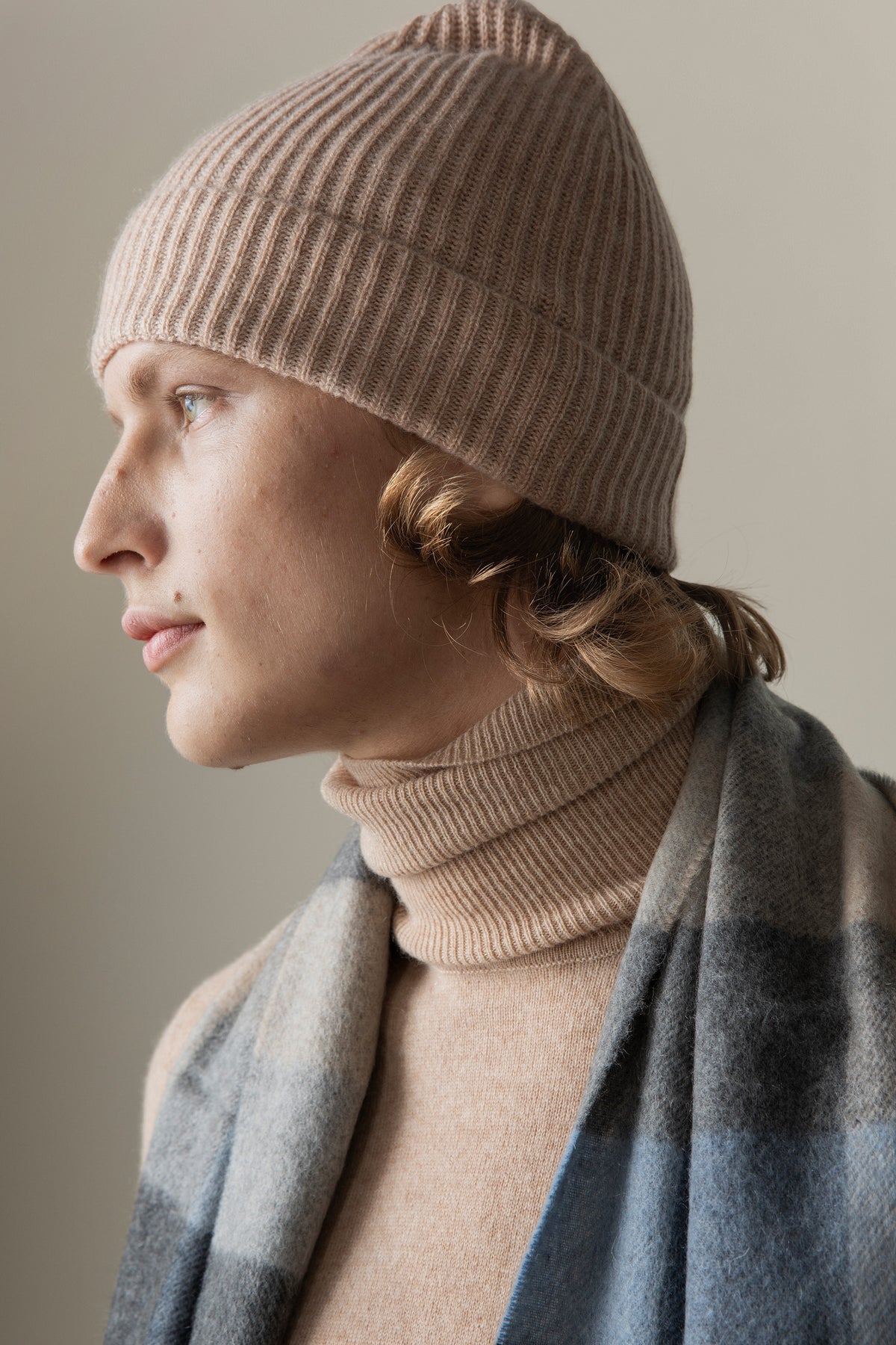 Unisex Cashmere Ribbed Beanie Hat in Sand worn by model in profile view, paired with matching turtleneck and plaid scarf