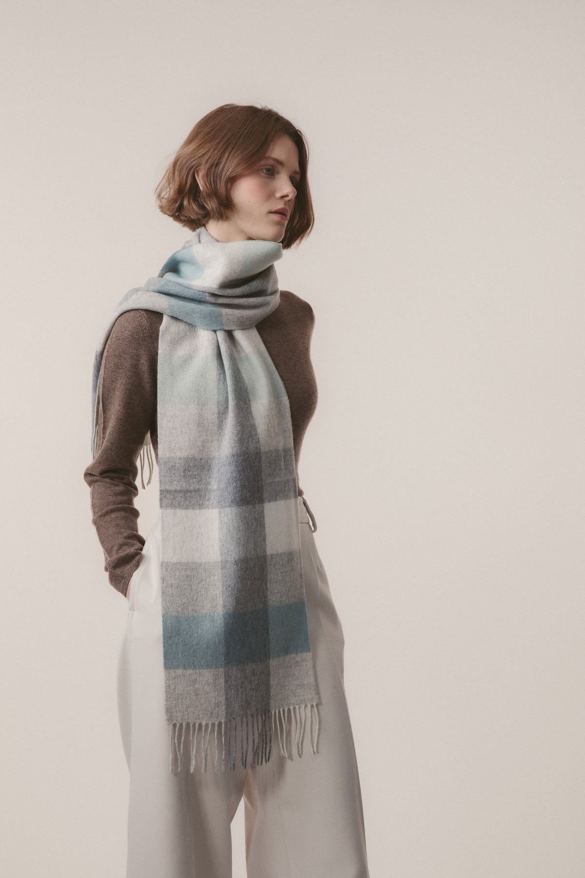 3 Square Check Lambswool Scarf in Sea Breeze worn wrapped around the neck, showing soft blue, grey, and white check pattern with fringed ends