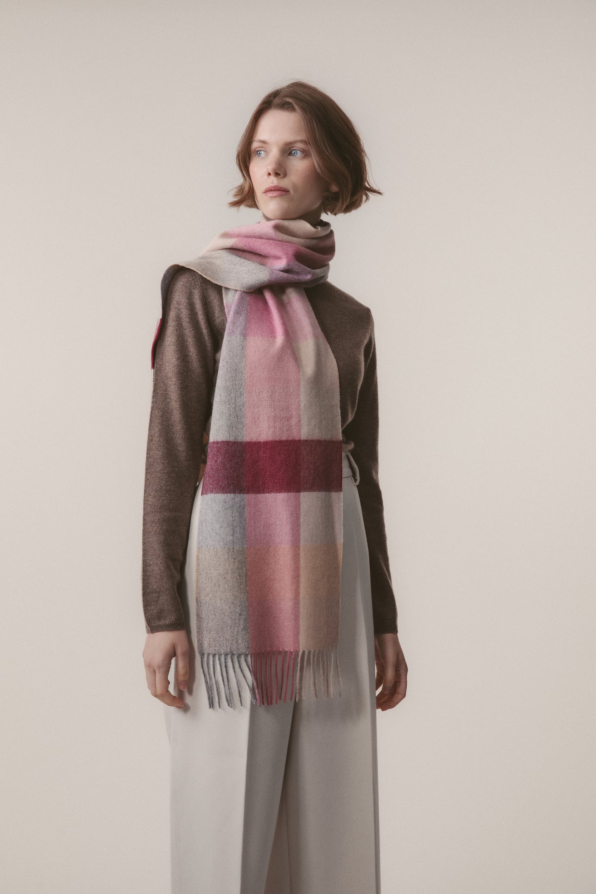 3 Square Check Lambswool Scarf in Summer Fruits worn draped around the neck, featuring pink, grey, and burgundy check pattern with fringed ends