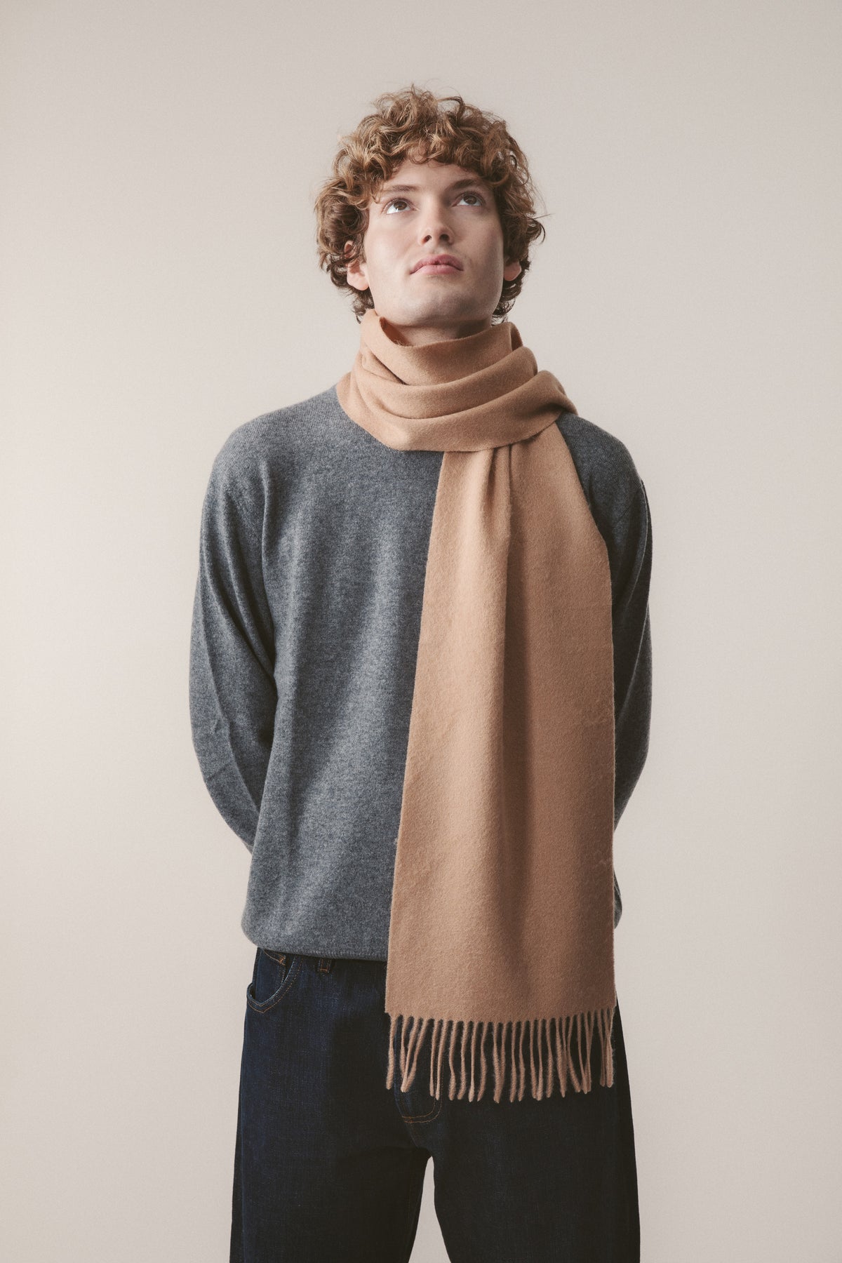 Solid Lambswool Scarf in Warm Camel worn looped around the neck by a model in a grey sweater, showing fringe detail