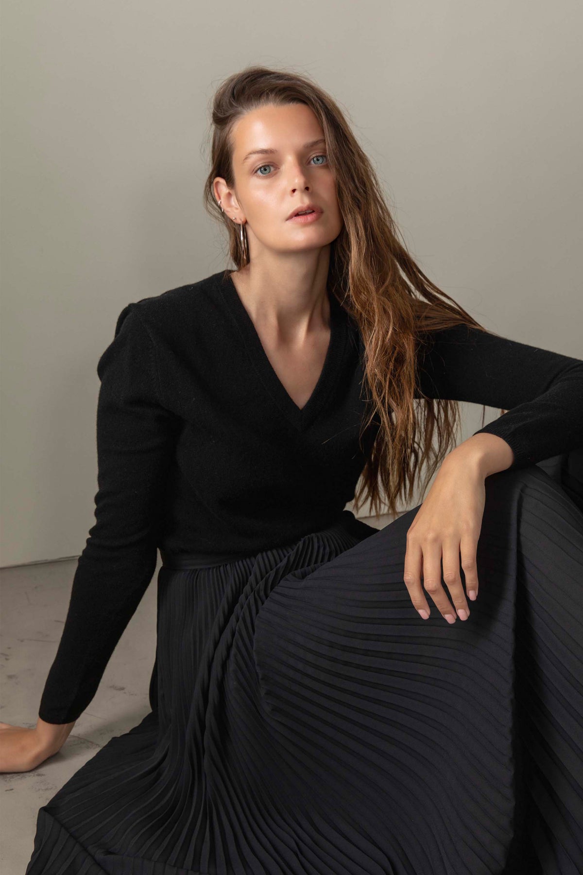 Model seated wearing Women's Cashmere V Neck Jumper in black, styled with a pleated skirt showing the relaxed fit and deep V neckline