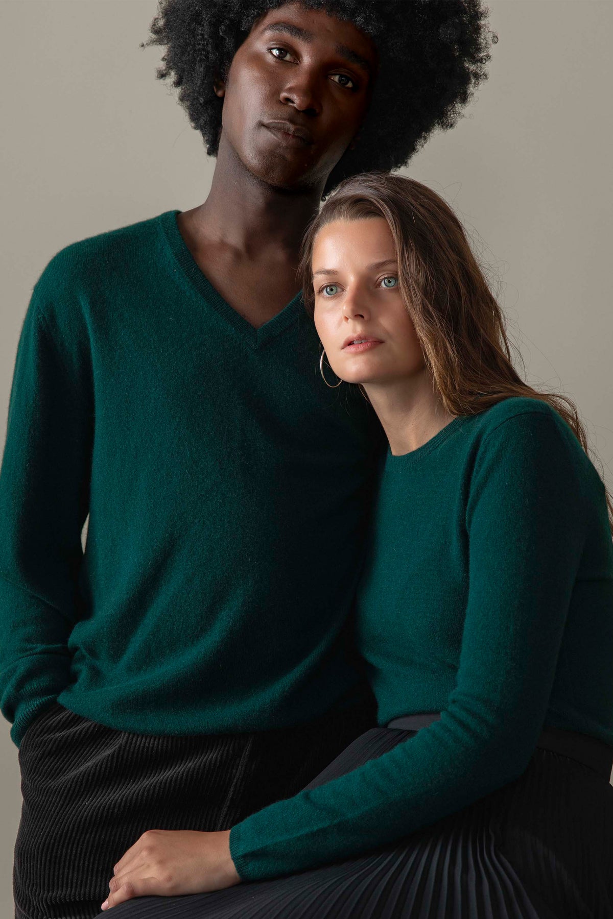 Two models wearing matching forest green cashmere jumpers — women's crew neck and men's v-neck styles shown together