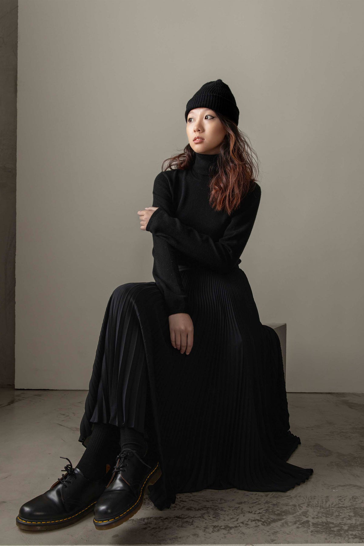 Women's Cashmere Polo Neck Jumper in black styled seated with pleated maxi skirt, beanie, and boots in all-black outfit