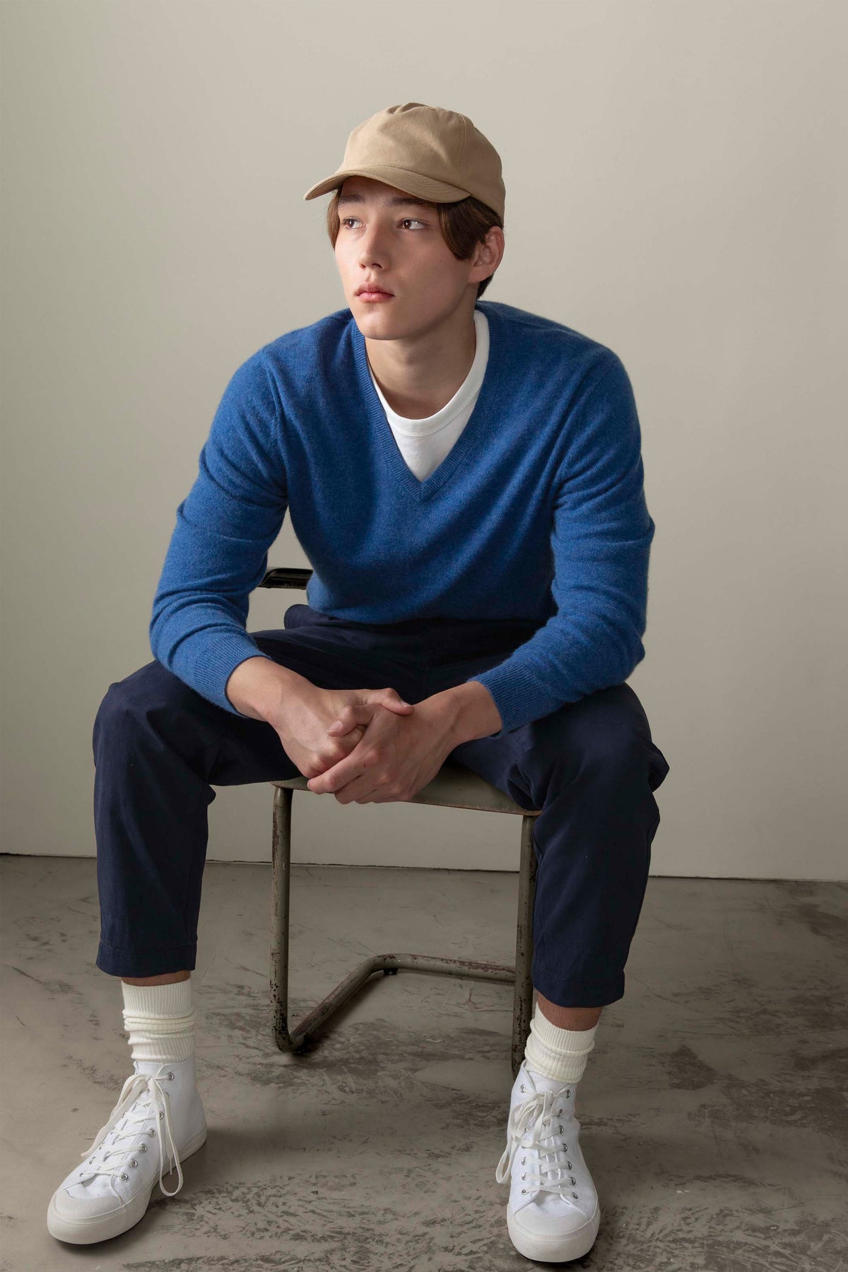 Model seated wearing Men's Cashmere V Neck Jumper in Denim blue over a white tee, styled with navy chinos and a tan cap
