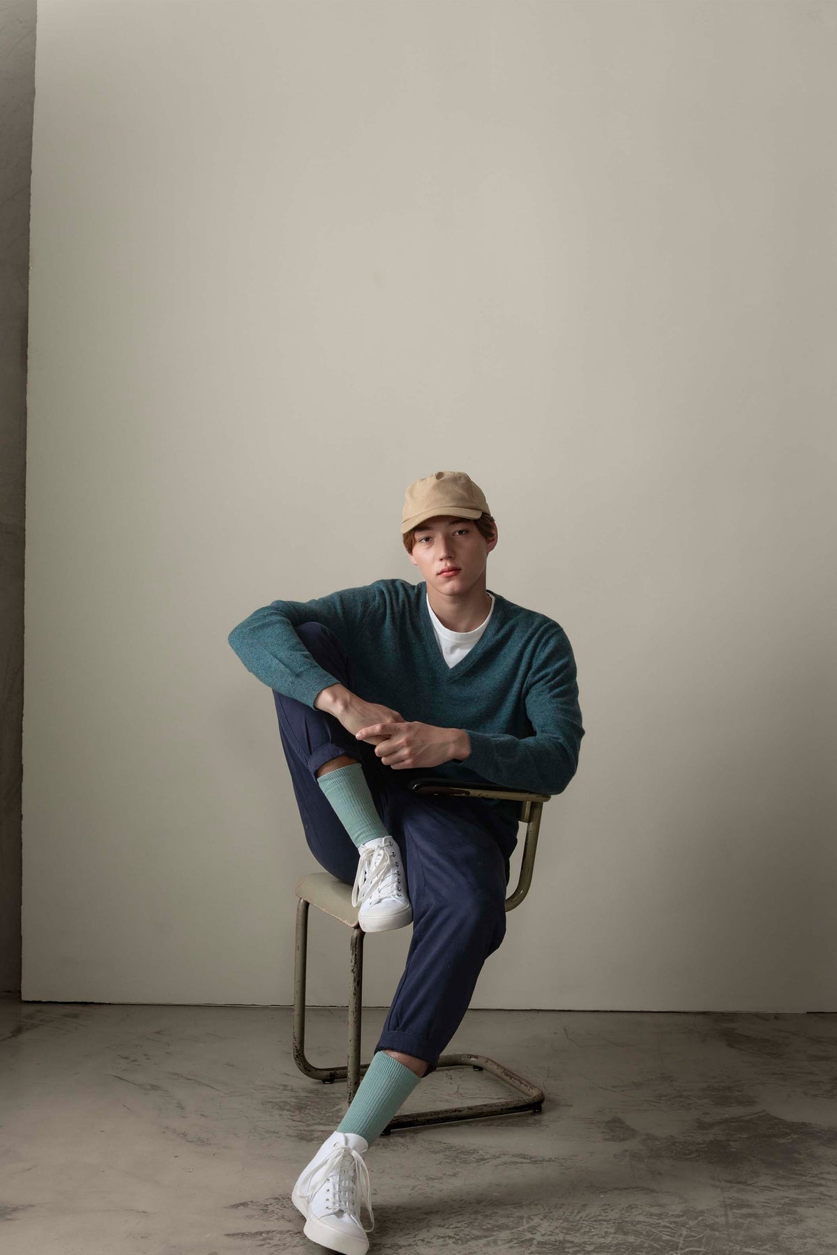 Model seated wearing Men's Cashmere V Neck Jumper in Haze Green over a white tee, paired with navy trousers and a tan cap