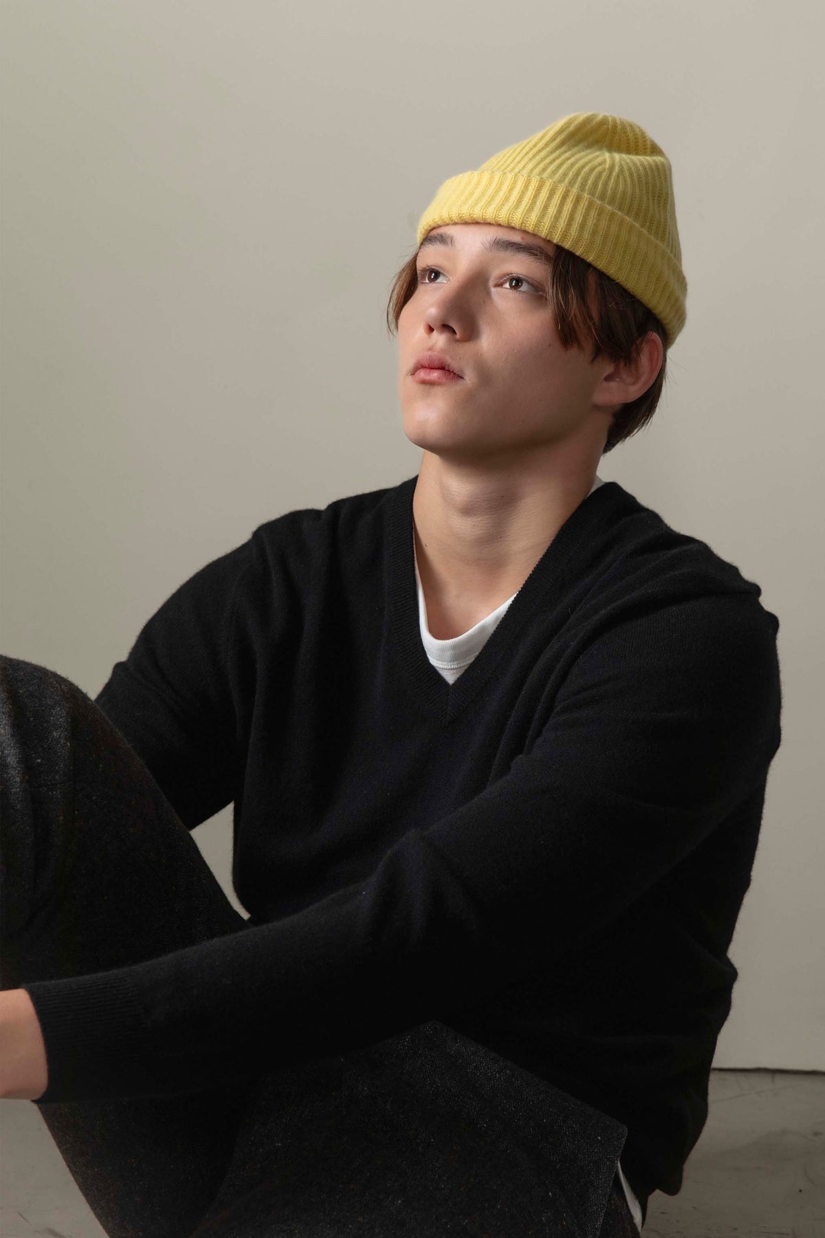 Model seated wearing Men's Cashmere V Neck Jumper in black, styled over a white tee with a yellow beanie