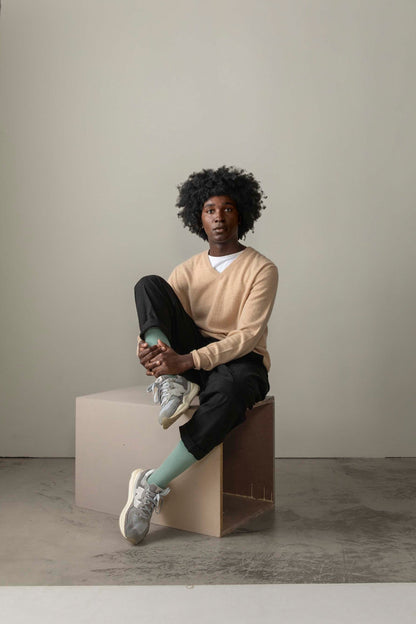 Model seated on a plywood cube wearing the Men's Cashmere V Neck Jumper in Vicuna over a white tee, paired with black trousers