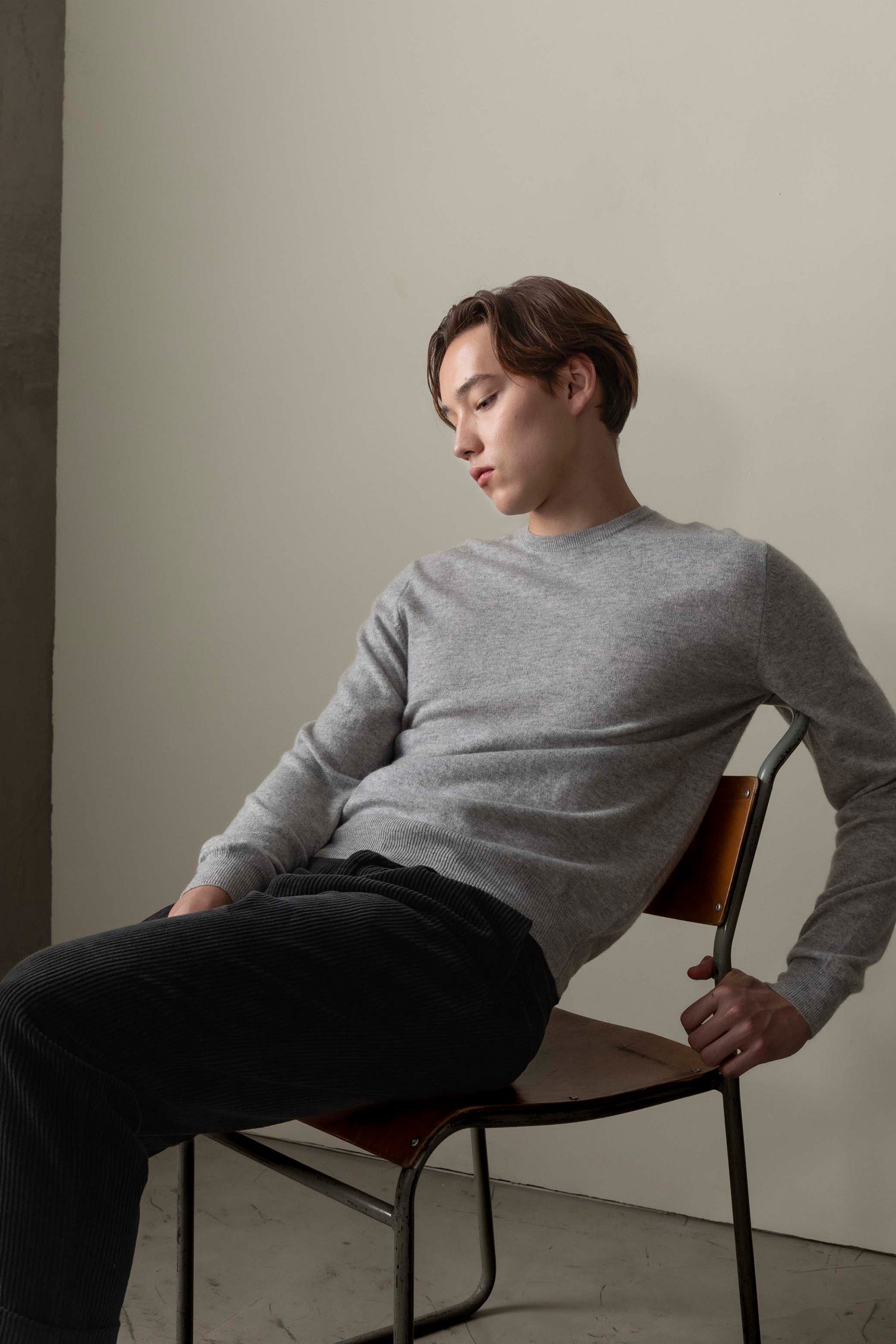 Model seated in chair wearing Men's Cashmere Crew Neck Jumper in light grey, showing relaxed fit and smooth knit texture