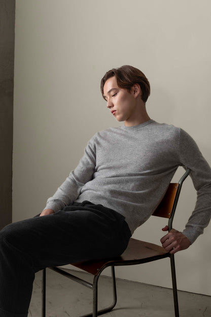 Model seated in chair wearing Men's Cashmere Crew Neck Jumper in light grey, showing relaxed fit and smooth knit texture