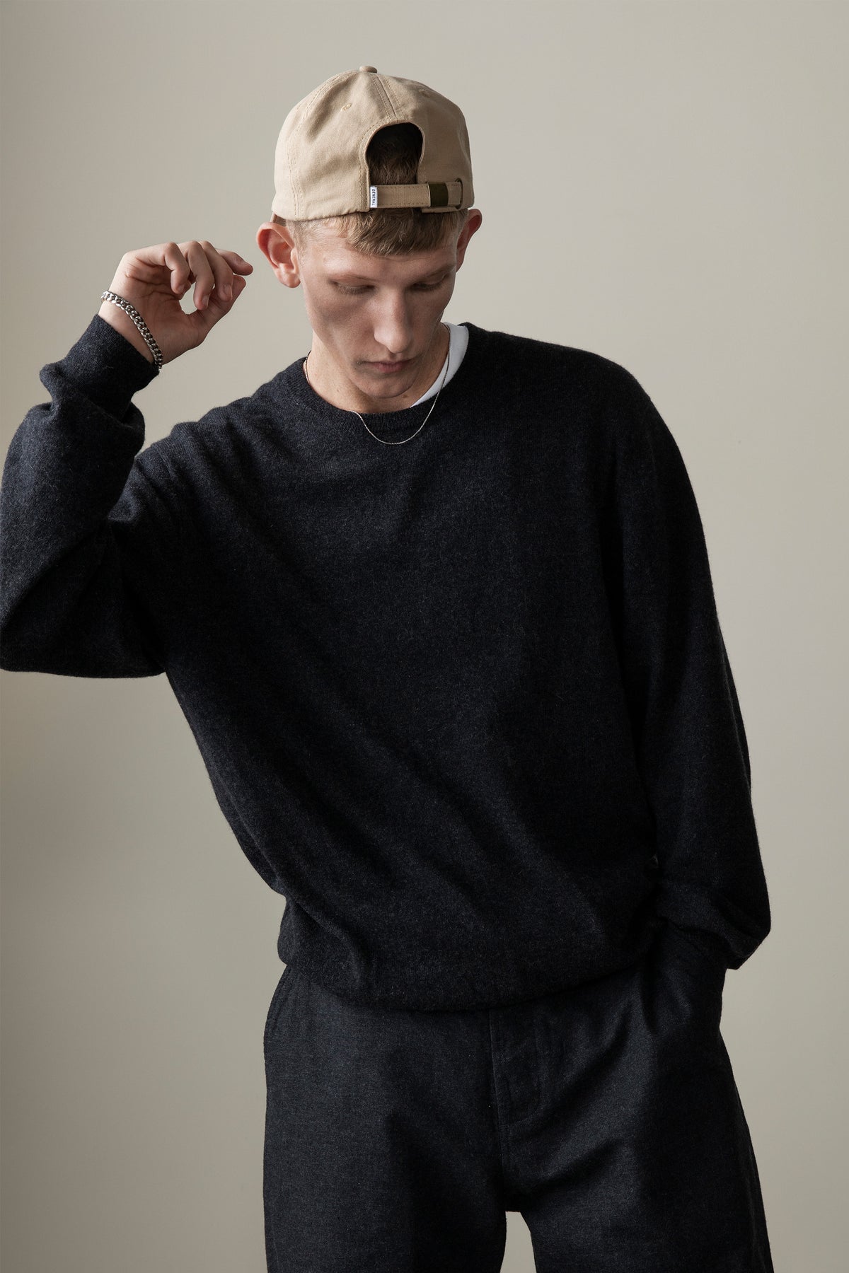 Model wearing Men's Cashmere Crew Neck Jumper in charcoal with relaxed oversized fit, styled with matching dark trousers and a tan cap