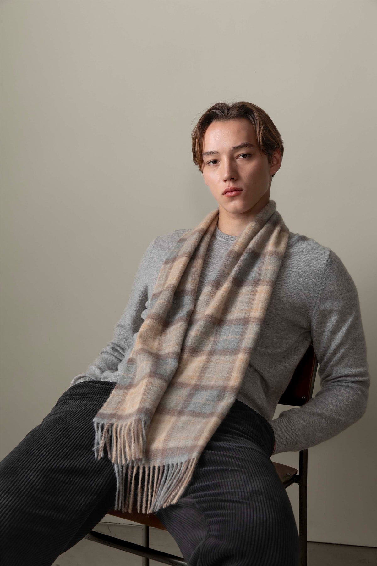 Model wearing the Made In Scotland Heritage Cashmere Scarf in Mackellar Natural draped over a grey sweater, showing plaid pattern and fringe detail