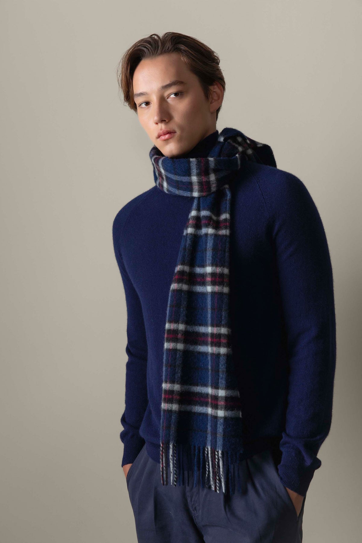 Model wearing the Scotty Thompson Cashmere Scarf in Navy, showing the blue tartan plaid pattern with white and red accents and fringed ends