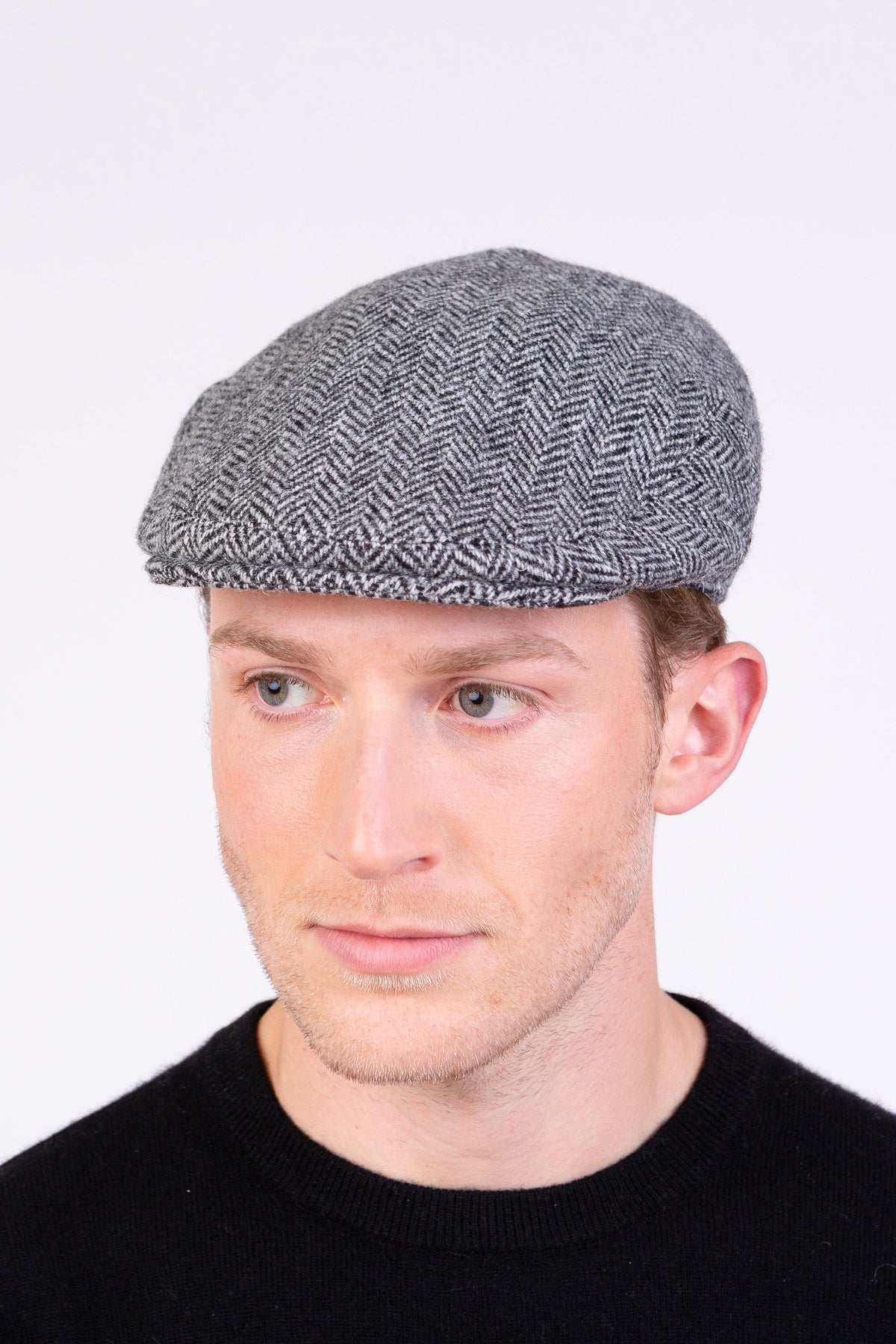 Harris Tweed Stornoway Cap in light grey herringbone wool, flat cap style worn by a male model, front-angled view