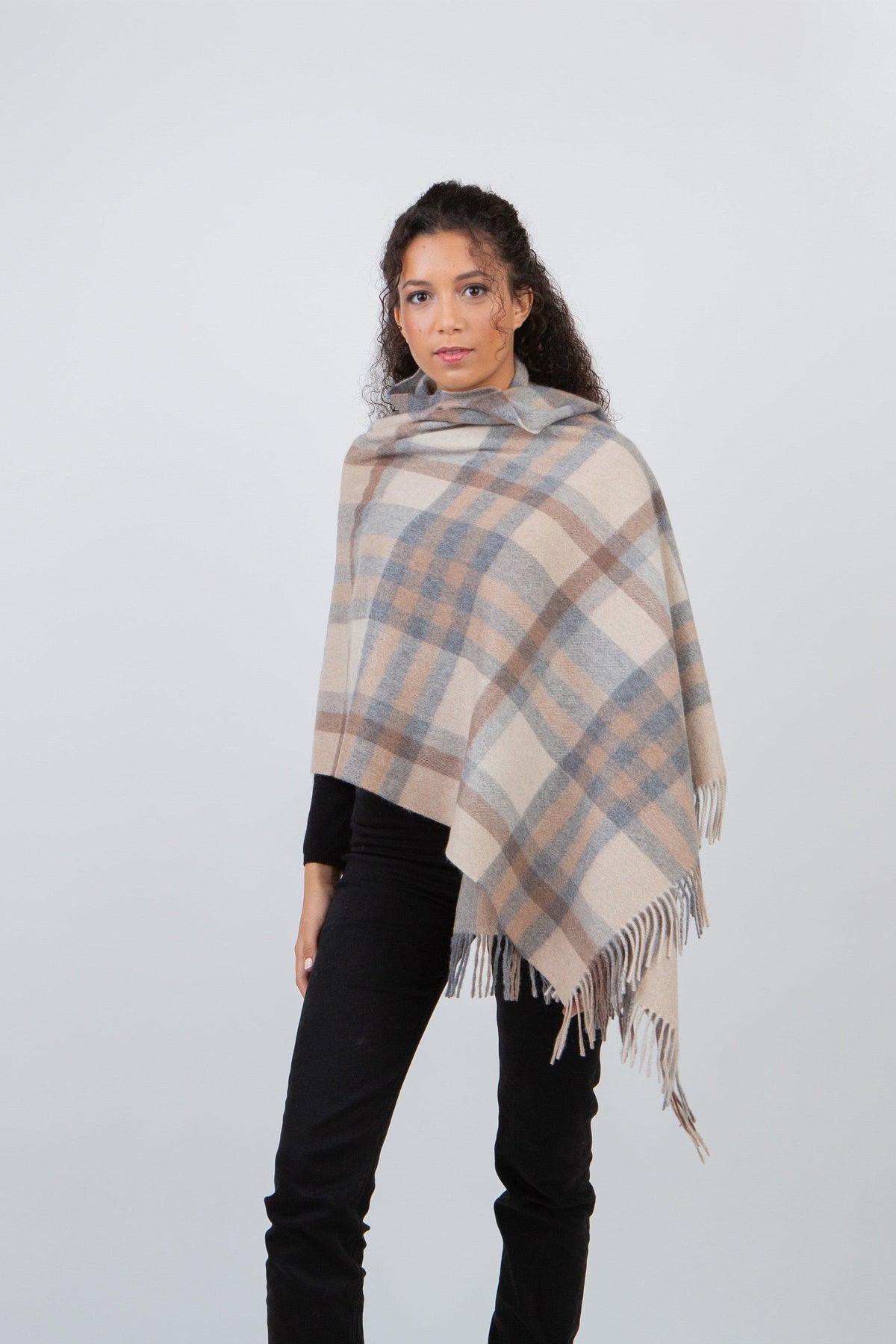 Model wearing Tartan 2 Lambswool Stole in Natural draped as a poncho, showing beige, grey, and brown plaid pattern with fringe hem