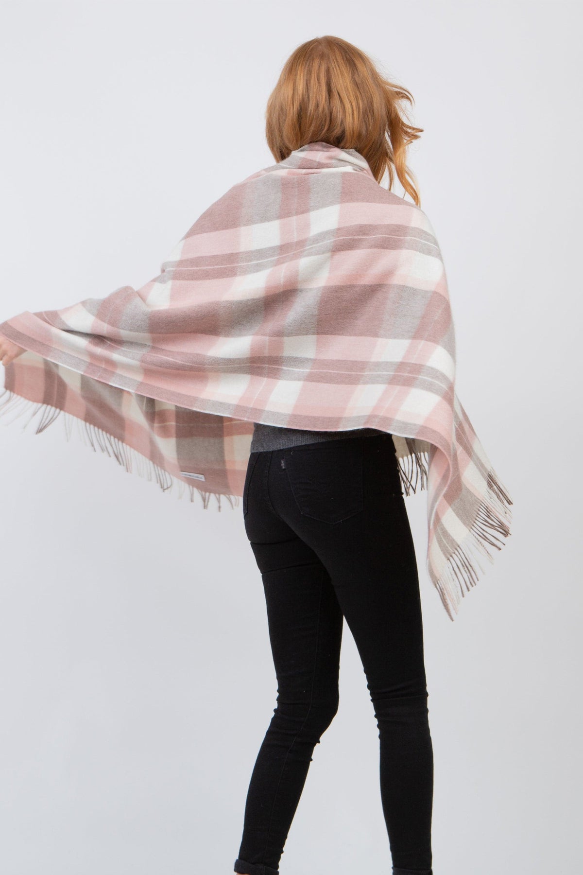 Back view of model wearing Contemporary Check Lambswool Stole in Cameo, showing pink, grey, and white plaid pattern with fringed edges