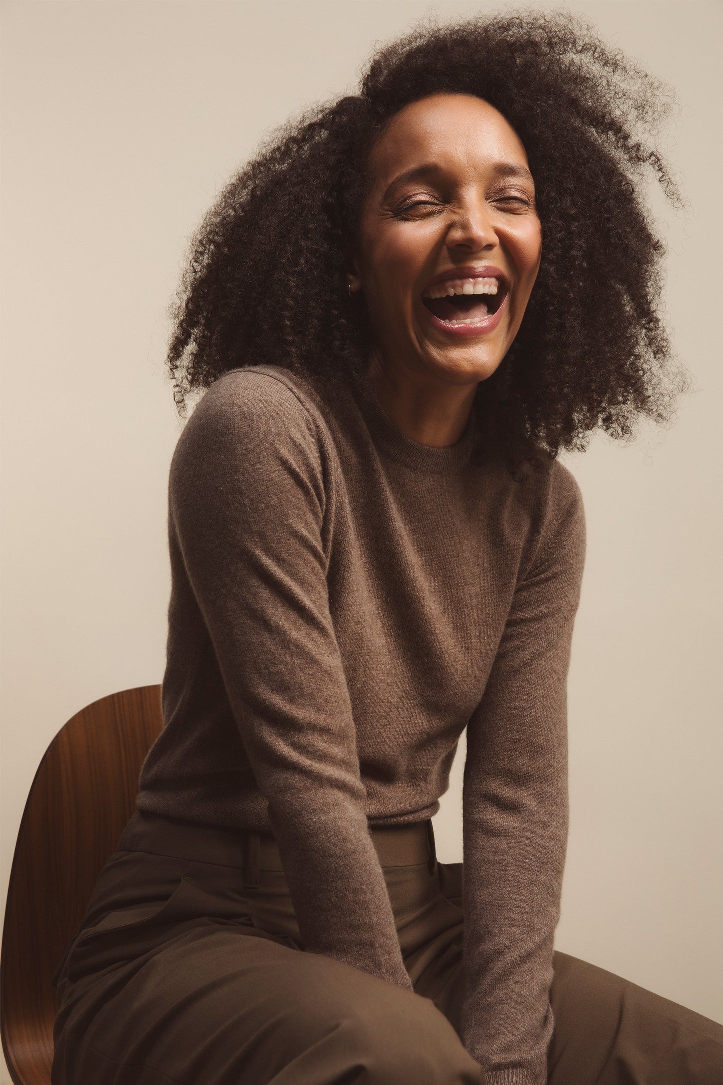 Model laughing while seated, wearing the Women's Cashmere Crew Neck Jumper in Squirrel brown, showing the soft knit texture and relaxed fit